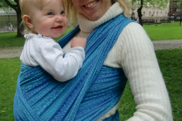 Bära en nyfödd bebis i bärsjal. Bebis ser väldigt glad ut i blå bärsjal, utomhus, sommar.