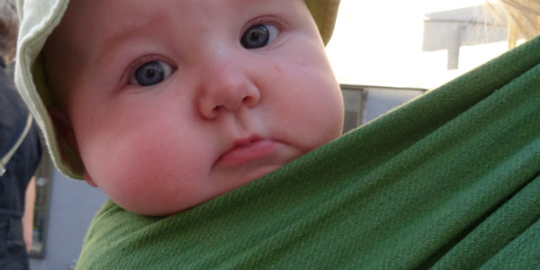 Bära en nyfödd bebis i bärsjal. Bebis i grön bärsjal tittar mot kameran med stora ögon.