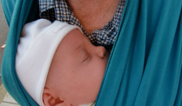 Bära en nyfödd bebis i bärsjal. Bebis sover i blå bärsjal på sin pappas bröst.