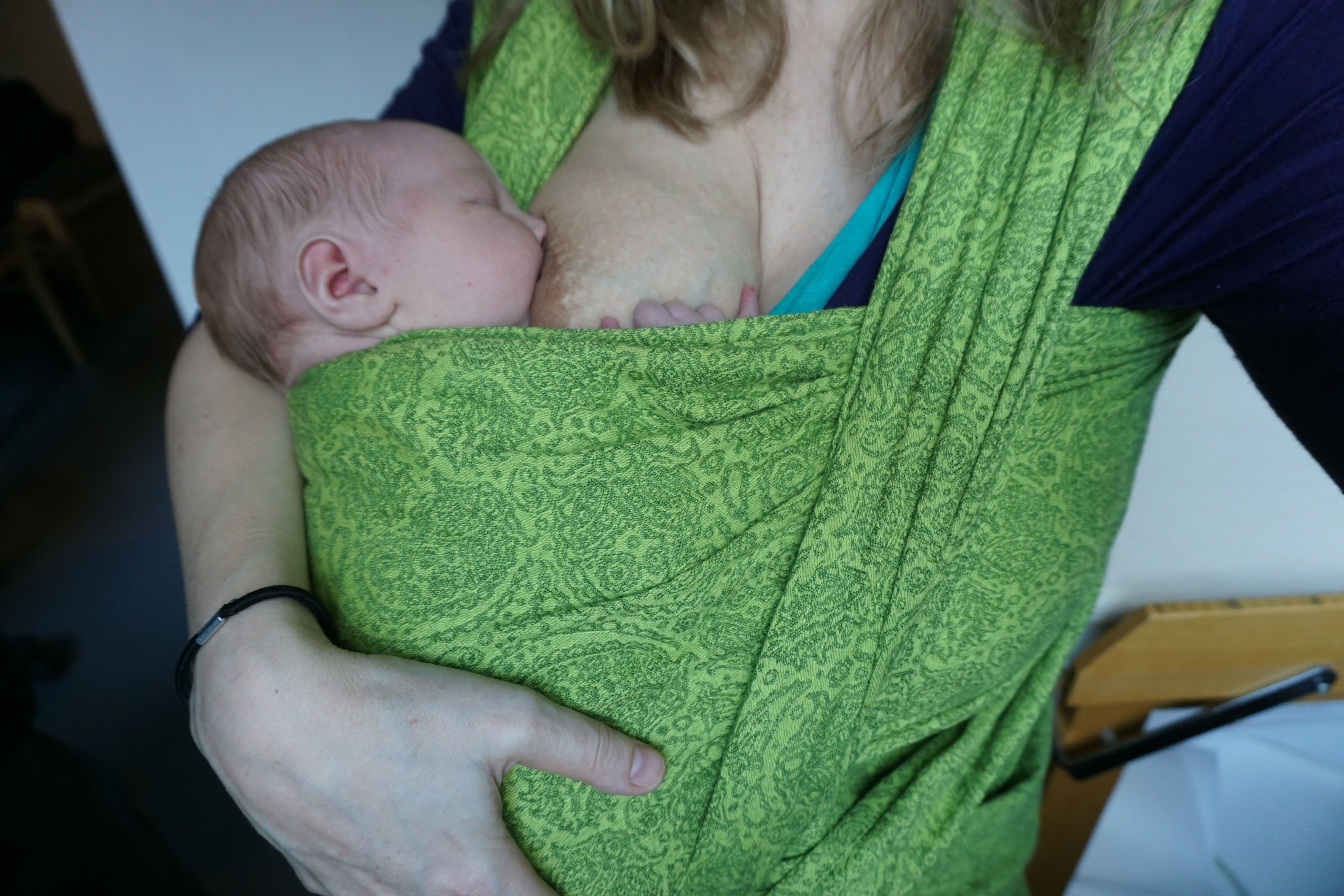 Bära en nyfödd bebis i bärsjal. Amning i grön bärsjal av tre dagar gammal bebis. En arm ger stöd för nacken.