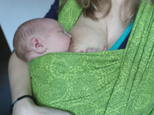 Nyfödd sväljer bröstmjölk. Med bebis i stadigt grepp i grön bärsjal.