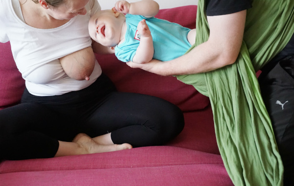 Amningsstrejk. Mamma har blottat bröst, sitter i soffan och får bebis langad till sig av pappa som burit bebisen i bärsjal. Bebisen skrattar.