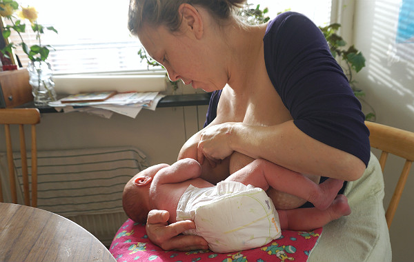 Stressen i förlossningsvården, amning. Mamma ammar bebis på stol i köket.