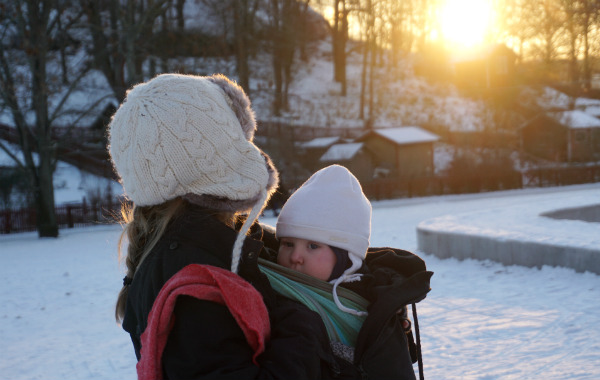 Amma i bärsele på vintern. Bebis ammar sittande upp utomhus, snö och låg sol.