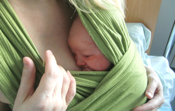 Bärsjal på BB. Alldeles nyfödd bebis sitter i grön bärsjal på mammans bröst, på BB-säng.
