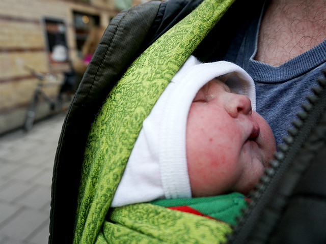 Stressen i förlossningsvården, amning. Nyfödd bebis i grön bärsjal på sin pappas bröstkorg, utomhus.
