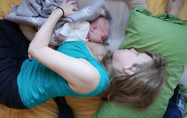 Liggamning, mamma och bebis ligger på sidan i säng. Bebis ligger i ett babynest och ammar.