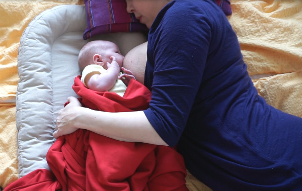 när du snart ska börja amma. Liten bebis ammar i babynest, mamman ligger på sidan på sängen.