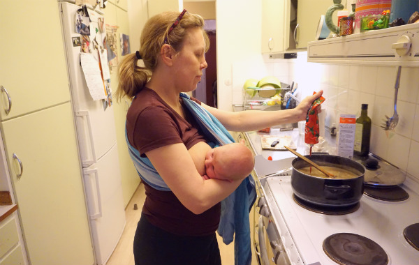 Amning i ringsjal. Kvinna ammar liten bebis i ringsjal, en typ av bärsjal, framför spisen.