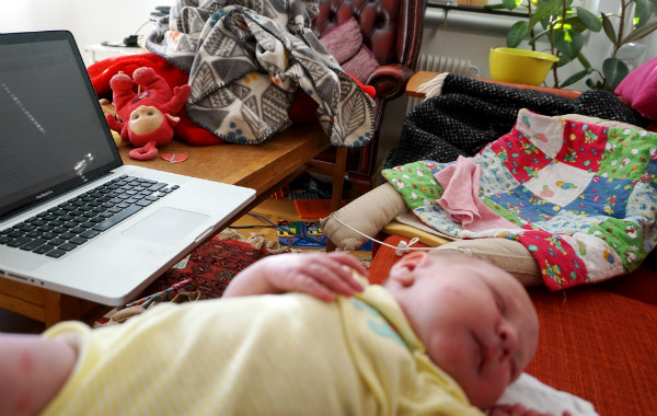 Amningsdagbok del två. Nyfödd bebis sover i förgrunden. Dator, stök och leksaker i bakgrunden, hemmiljö.