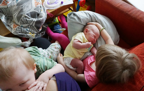 Amningsdagbok del fyra. Tre barn i soffan, tvååring vid mammas bröst, nyfödd får klapp på huvudet av storasyster.