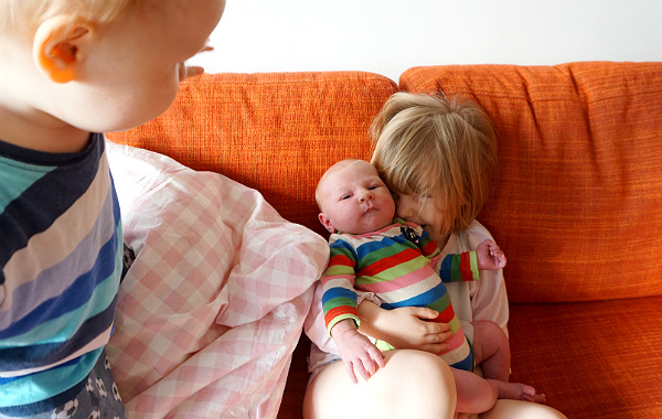 Amningsdagbok del tre. Nyfödd bebis i knät på storasyster i soffa, storebror ser på.