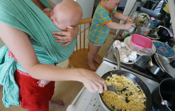 Tips om hur du ammar i bärsele och bärsjal. Bebis ammar i grön bärsjal medan mamman steker ägg. Lite äldre barn i bakgrunden, kök.