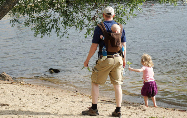 Bära barn på ryggen, pappa på strand med fyraåring som går i sanden samt bebis i bärsele på ryggen.