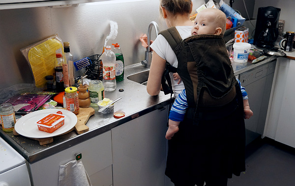 Bära barn på ryggen. Bebis i bärsele på mammas rygg i köket, mamma diskar.