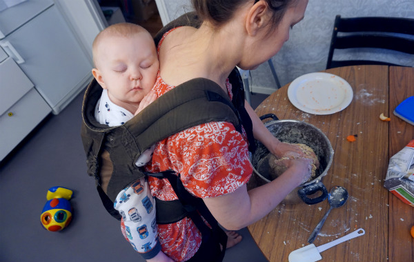 Bära barn på ryggen. Bebis sover i bärsele på mammas rygg, mamma bakar, kök.