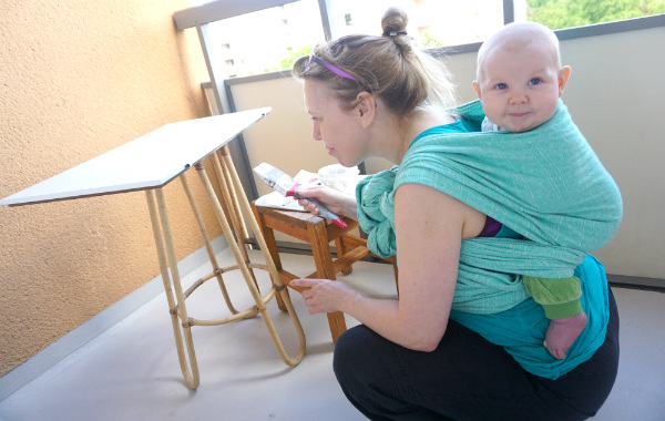 Bära barn på ryggen. Bebis i bärsjal på mammas rygg på balkong, mamma sitter på huk och målar.