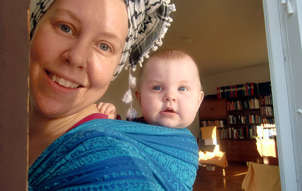 Bära barn på ryggen. Bebis i bärsjal på mammans rygg. Mamman har en sjal om huvudet.