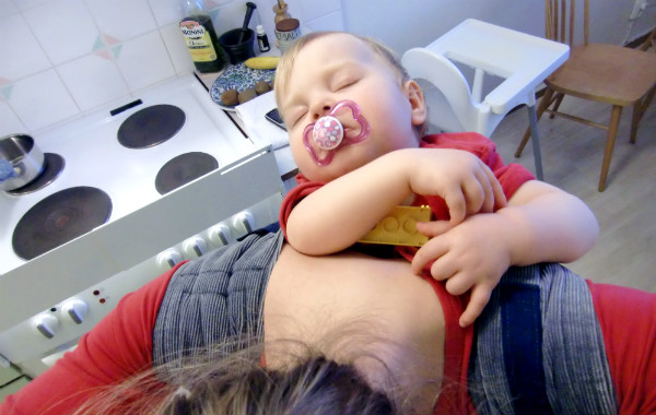 Bära barn på ryggen. Bebis har somnat i beh dai med en duplo-bit i handen, kök.