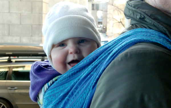 Mest förvirrande sakerna med amning. Glad bebis i bärsjal på pappas rygg, vinter, stadsmiljö.