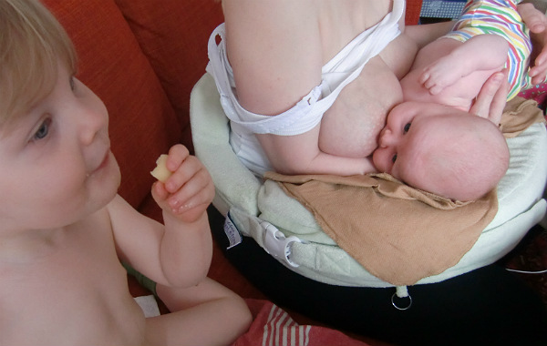 Tilläggsmata mindre, ge mindre ersättning. Liten bebis ammar på amningskudde i soffa, äldre syskon sitter bredvid och äter något.