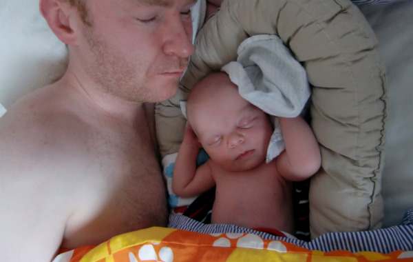 Pappa sover bredvid nyfödd bebis, som ligger i babynest.