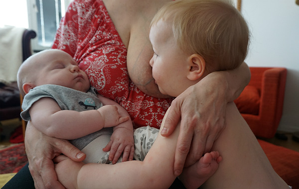 Tvååring ammar medan bebis sover i mammans famn.