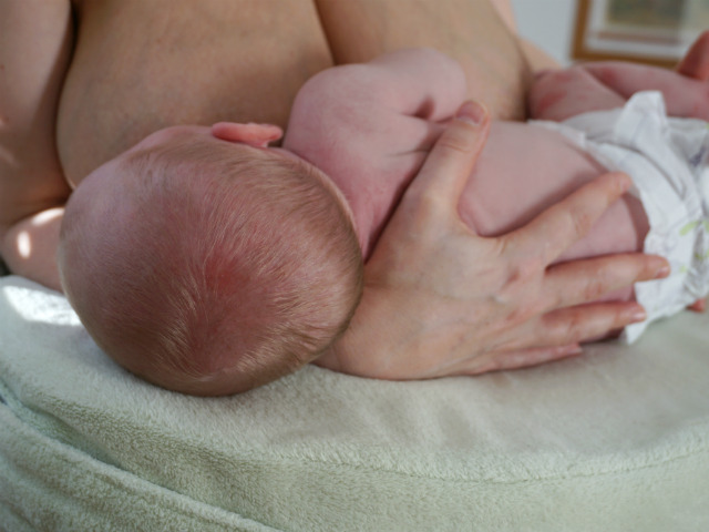 Amning och ont i ryggen. Bebis ammar på amningskudde, framifrån sett. Hand hålls om barnets rygg.