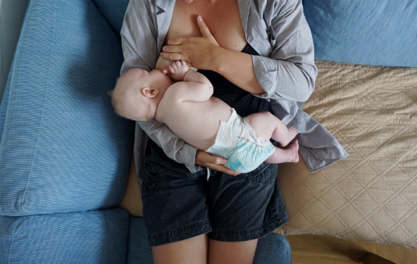 Amma max 15 minuter per bröst. Kvinna ammar bebis halvsittande på soffa.