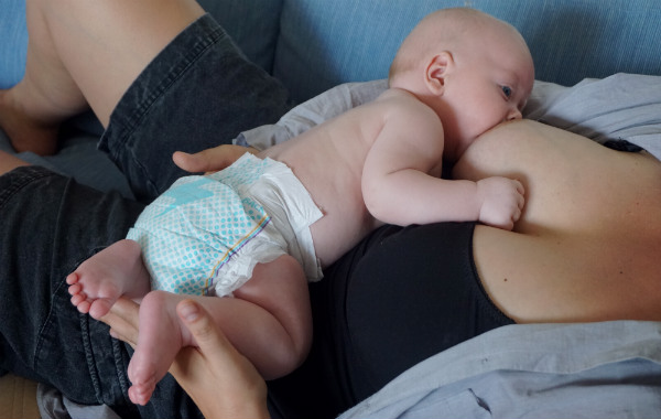 Amma max 15 minuter per bröst. Bebis i bara blöjan ligger ned och ammar på sin mamma, som ligger på rygg.