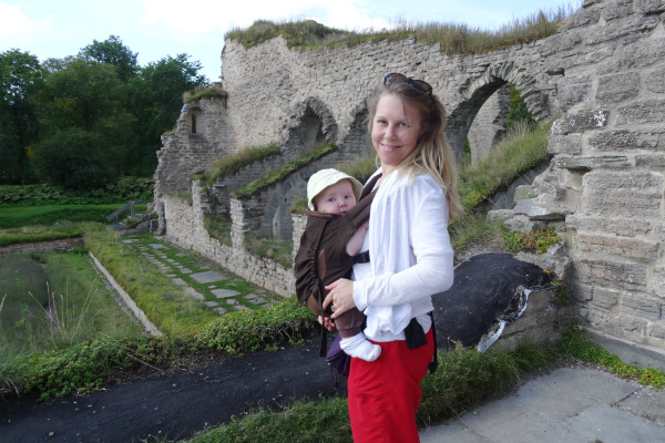 Amning och bära barn. Barn i brun bärsele, mamma och barn framför ruin, sommar.