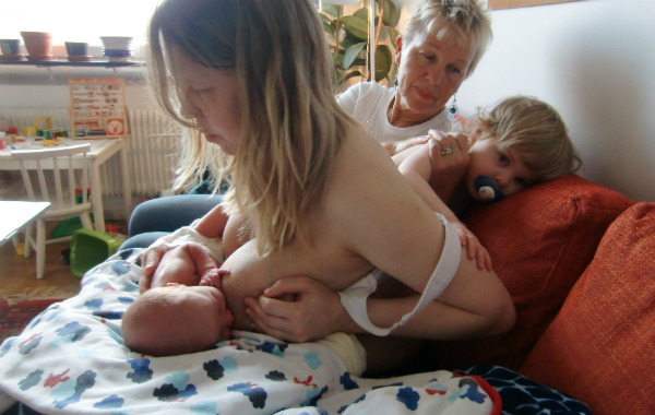 Amning och ont i ryggen. Kvinna ammar framåtlutad i soffa. I bakgrunden mormor samt äldre syskon till bebisen.