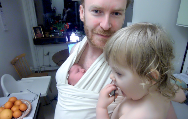 Tilläggsmata mindre, ge mindre ersättning. Pappa bär nyfödd bebis i vit bärsjal, har större syskon på sin arm samtidigt.
