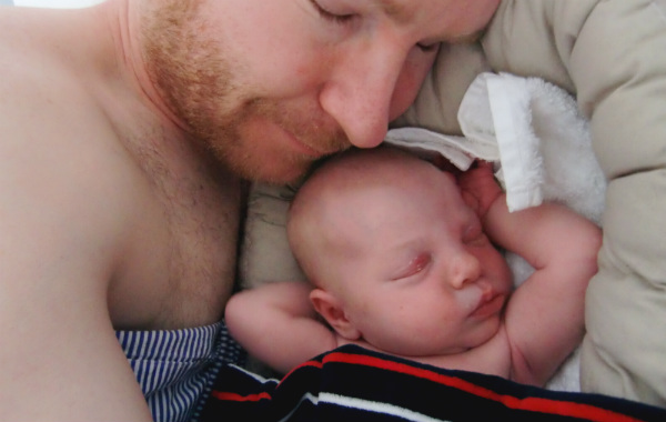 Tilläggsmata mindre, ge mindre ersättning. Pappa gosar med nyfödd bebis som sover i babynest.