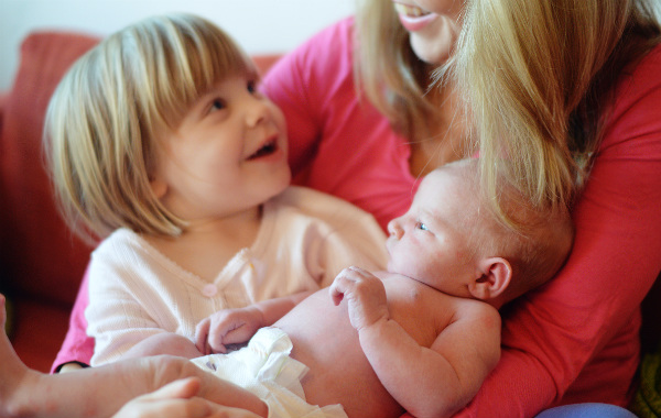 Mest förvirrande sakerna med amning. Inomhus, nyfödd bebis i famnen på mamma, större syskon sitter också i famnen och tittar leende upp mot sin mamma.