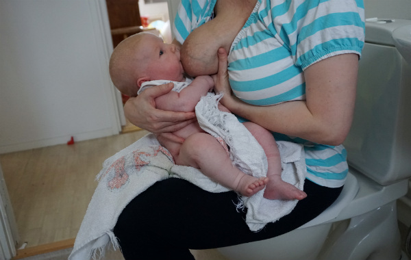 Mest förvirrande sakerna med amning. Nyfödd bebis ammar halvsittande med mamma sittande på toalettlock.