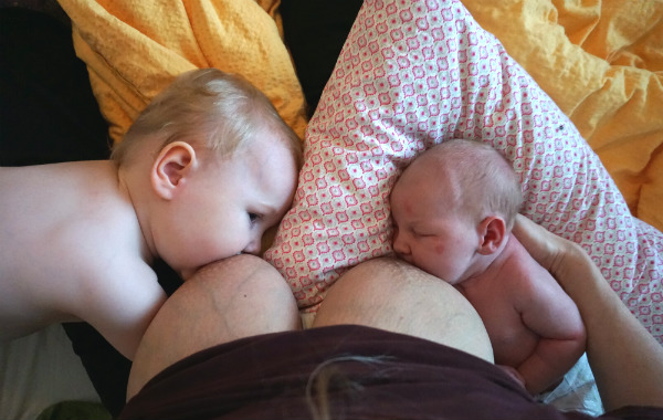 Tandemamning. Bebis och större syskon ammar samtidigt. Båda ligger ned, från varsitt håll, ovanpå säng. Mamman sitter upp.