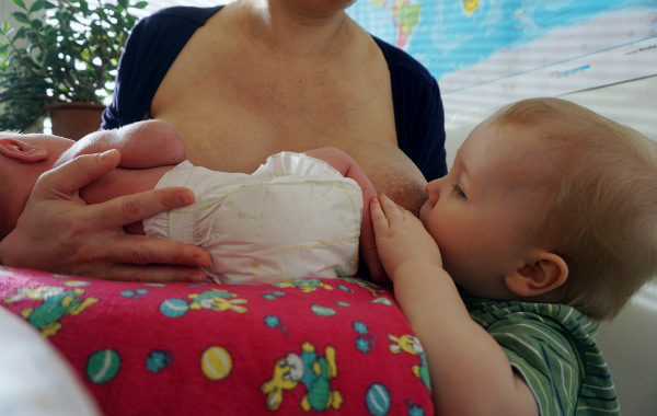 Tandemamning. Större syskon och bebis ammar samtidigt. Det större syskonet står upp, det mindre ligger på kudde. Mamma sitter på stol.