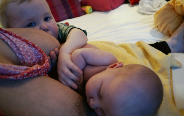 Tandemamning. Bebis och större syskon har ammat, bebisen har somnat vid bröstet. Syskonen håller varandra i handen.