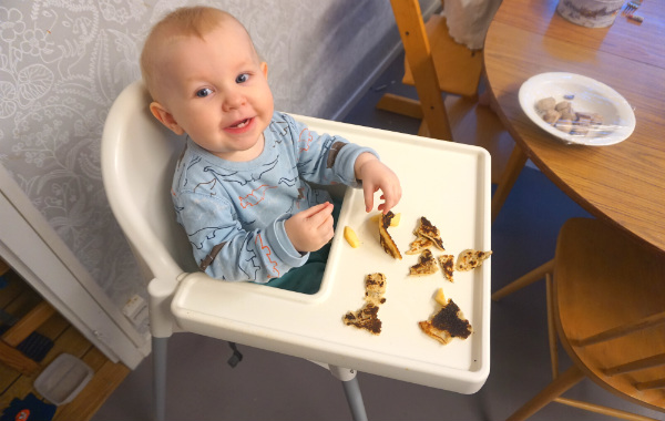 Ettårigt barn äter en pannkaka, i bitar, i matstol.