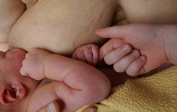Amningsbildextra. Äldre barn sträcker fram handen mot ammande nyfödd bebis hand.