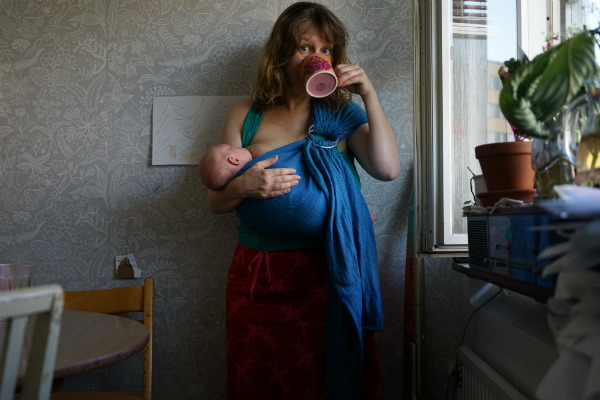 Amningsbildextra. Kvinna ammar barn i bärsjal, stående i kök med kopp vid munnen.