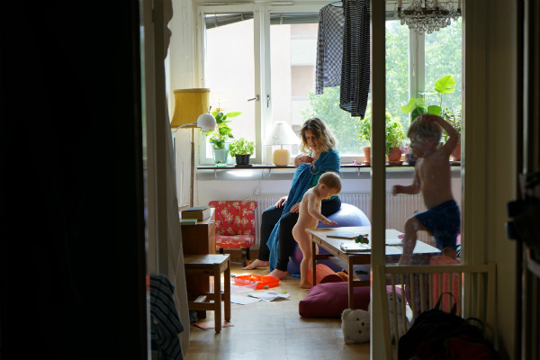 Amningsbildextra. Kvinna med bebis i bärsjal på pilatesboll i vardagsrum, två äldre barn springer omkring.