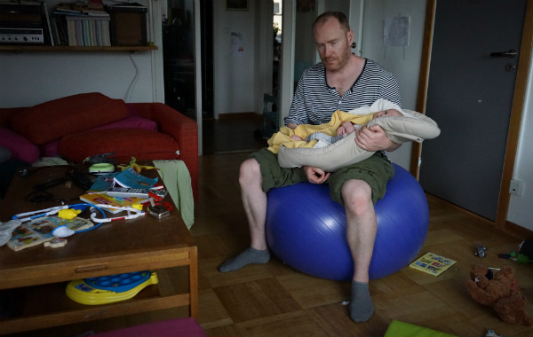 Amningsbildextra. Man vaggar bebis i babynest på pilatesboll i vardagsrum.