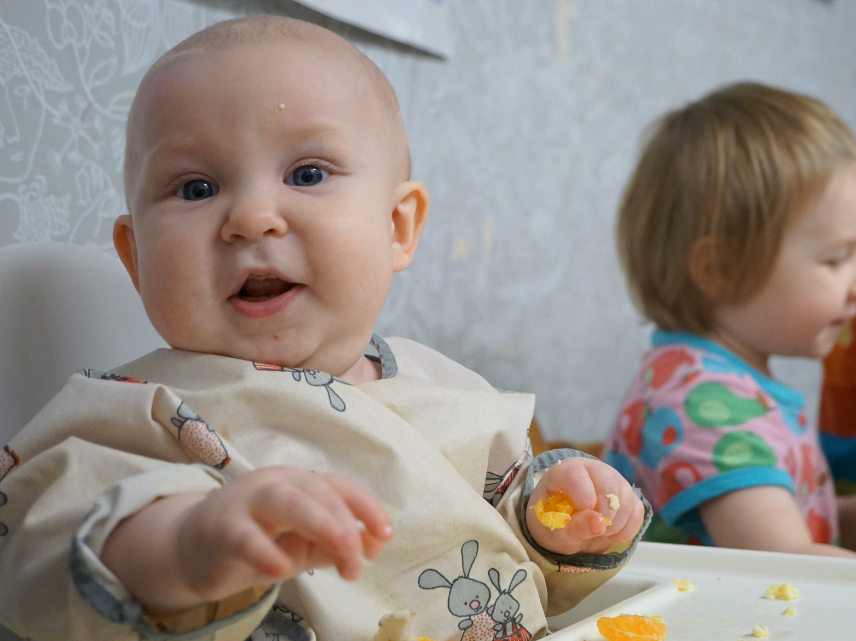 Matintroduktion, BLW. Bebis i förkläde äter själv, i stol i kök. Syskon skymtar i bakgrunden.